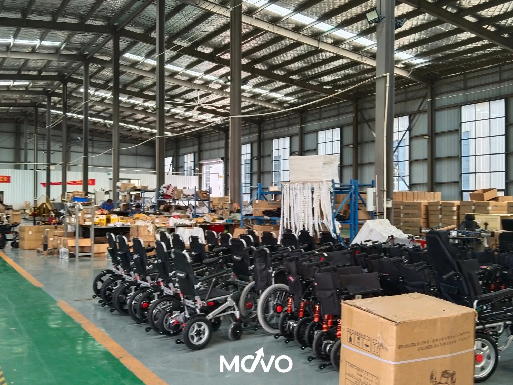 Movo Chair factory floor — rows of chairs staged for assembly under wide industrial skylights, technicians working at benches in the background, cardboard shipping cartons stacked along the wall, MOVO watermark centered on the image