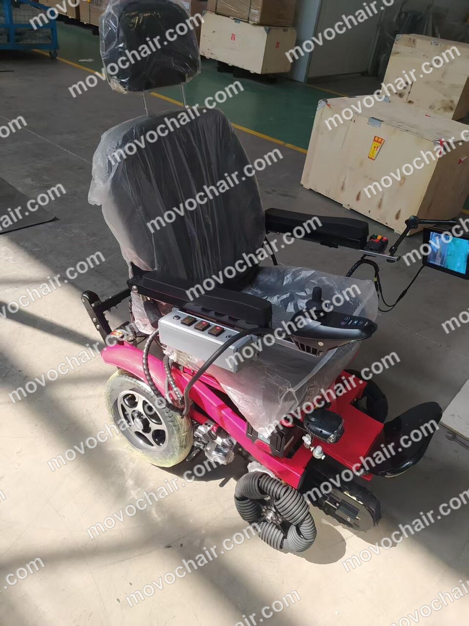Movo X on the factory floor — pink frame, joystick controller and display installed, stair-climbing rubber tracks visible under the seat, protective plastic wrap still on